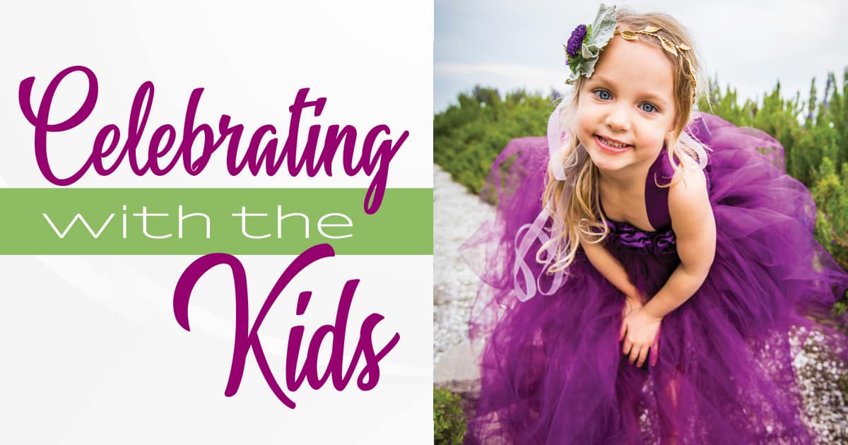 A smiling young girl in a purple tulle dress and flower crown stands on a sandy path beside green shrubs, with "Celebrating with the Kids" text.