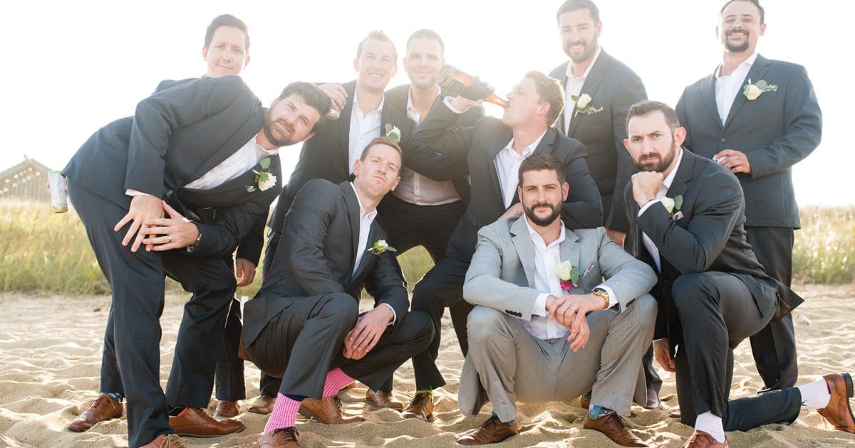 A group of groomsmen in dark suits and one light gray suit pose playfully on a sandy beach, one holding a bottle.