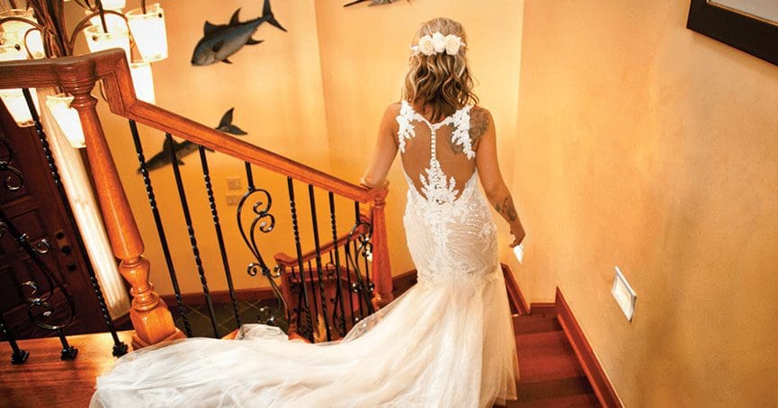 A bride in a lace fitted gown with a sheer back and floral hair flowers climbs a wooden staircase.