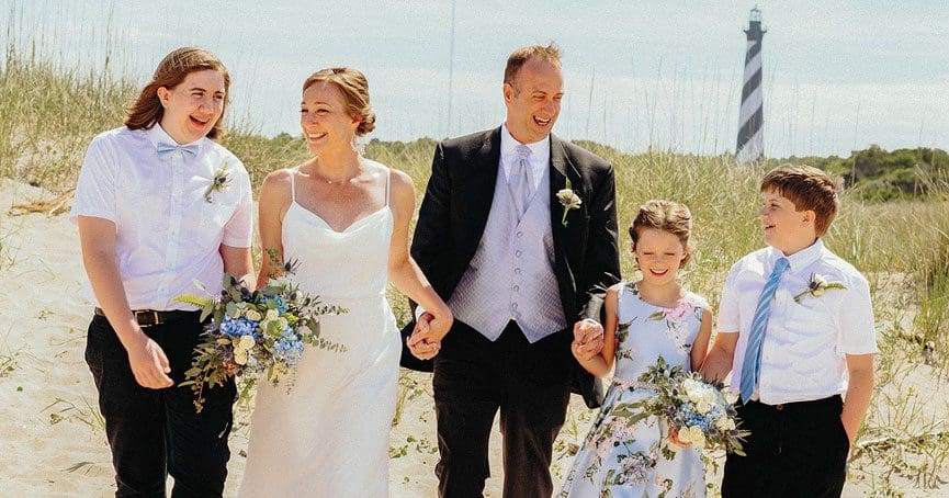A family of five walks through sea oats on a sandy dune, with a black-and-white striped lighthouse behind them.